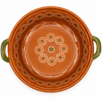 An overhead view of a round terracotta clay casserole dish with green handles and hand-painted white and green decorative patterns on the inside.