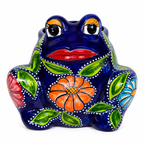 A ceramic Mexican-style painted frog piggy bank on a white background. The frog's body is glossy dark blue, decorated with hand-painted green leaves, a large orange flower on the front, a red flower on the side, and smaller blue and orange flowers.