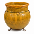A large decorative ceramic cauldron-shaped planter in a textured mustard yellow and ochre glaze, featuring embossed vertical panels, a patterned band around the neck, and a dark metal stand with three legs and side rings, shown on a white background.